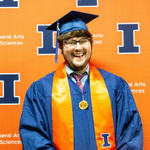 Portrait of Ian Garvey in graduation regalia against an orange background with Block I letters