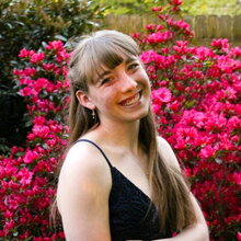 Outdoor portrait of Rachel Schaaf standing in front of a bright pink flowering bush.  