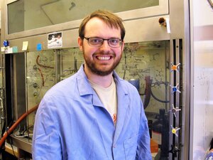 Andrew Zahrt stands in a blue lab coat in a chemistry lab with equipment in the background.