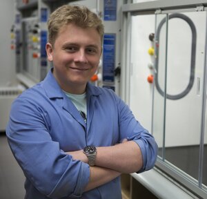 Portrait of Alex Shved in a blue lab coat with arms crossed standing in a lab on campus. 