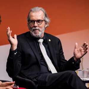 A photo of Ralph Nuzzo in a dark suit and light-colored tie in front of an orange-colored wall during a live interview in Norway.