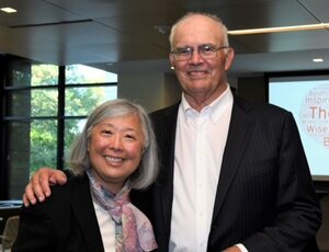 Susan Morisato, on left, stands next to Tom Remec in a room with a window and window panes behind them to the left and a screen behind them to the right.