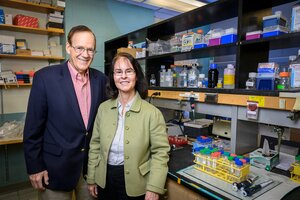 John Katzenellenbogen stands to the left of Benita Katzenellenbogen in a lab with shelves in the background.