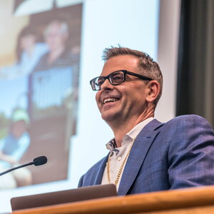 Burke standing at the podium smiling during his speech with a overhead screen in the background